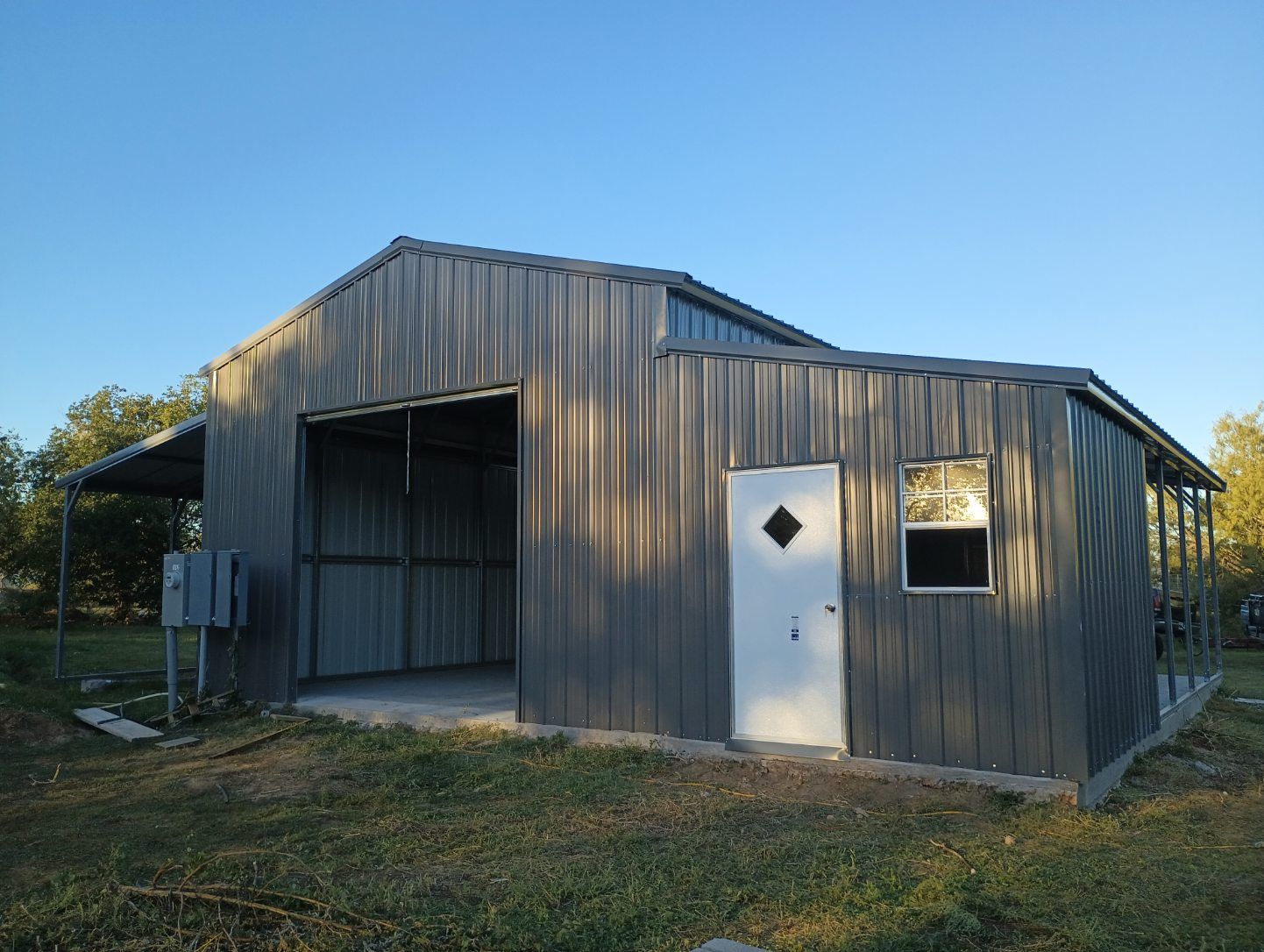 Gray metal shed with open bay door and white entry door in a grassy yard under a clear blue sky