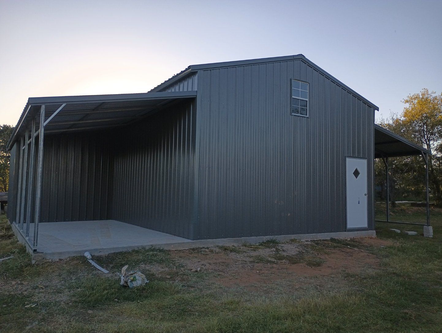 Metal barn with an open side shed at dusk, standing on a grassy lot.