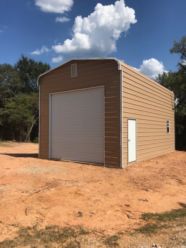 Tan metal storage building with roll-up door on red dirt under a blue sky with clouds