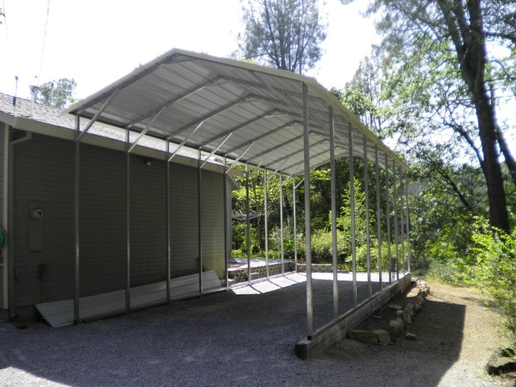 Covered metal carport beside a green building in a wooded outdoor area