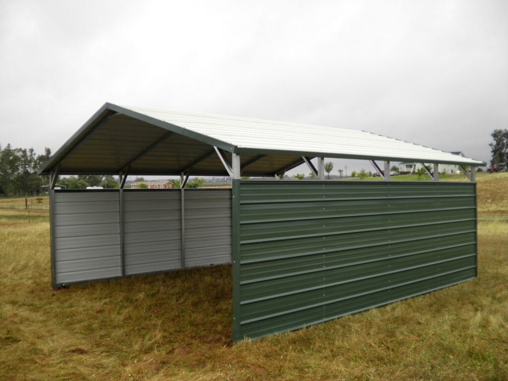 Metal livestock shelter with green and gray corrugated walls and a sloped roof in a grassy field