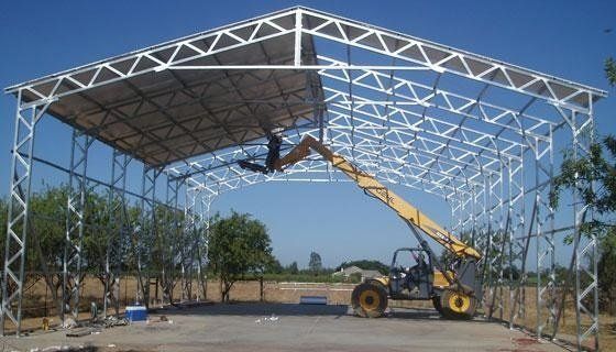 Construction site with a yellow crane lifting steel roof trusses on a large open-frame building.