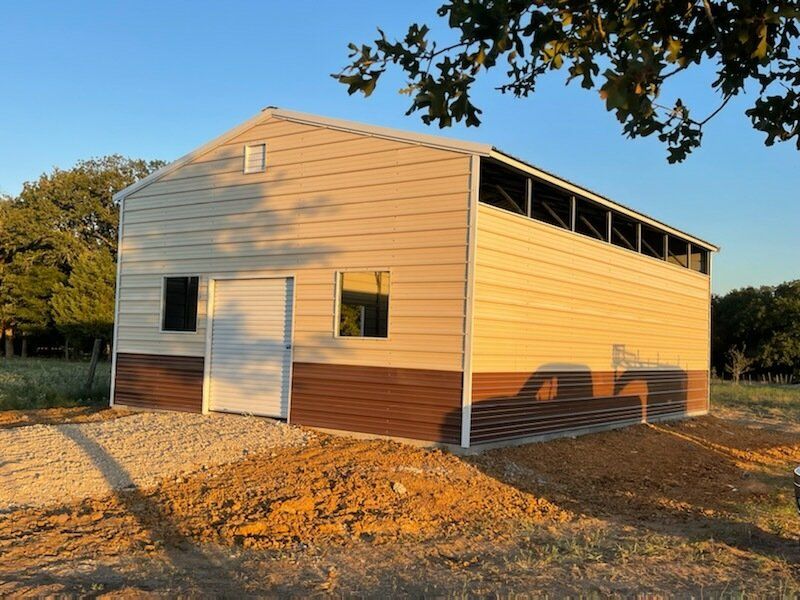Tan-sided barn with white trim and open side vents on a dirt lot at sunset