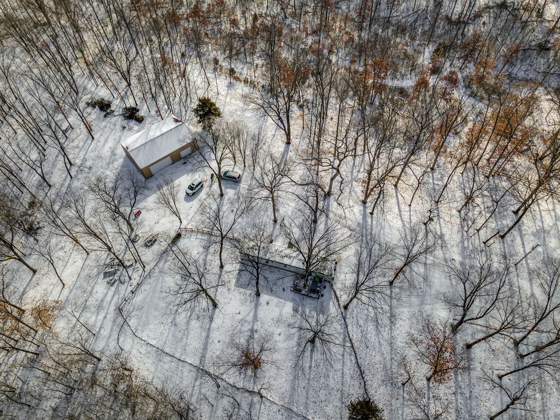 Aerial view of a snowy wooded area with two small buildings and leafless trees.