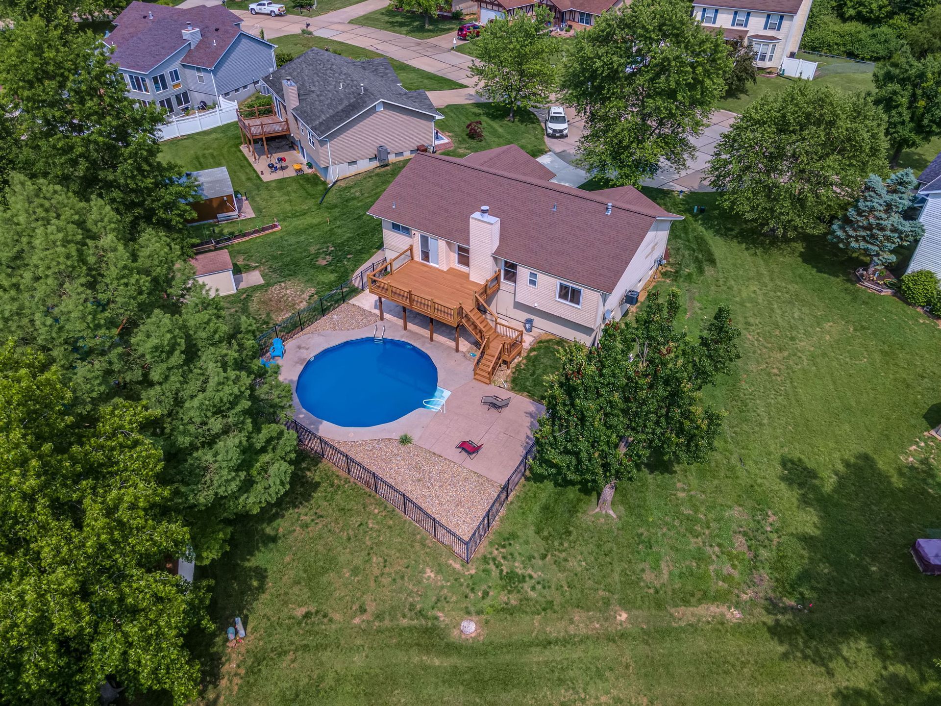 Aerial view of a suburban backyard with a blue round pool, patio, and beige house surrounded by green lawns and trees