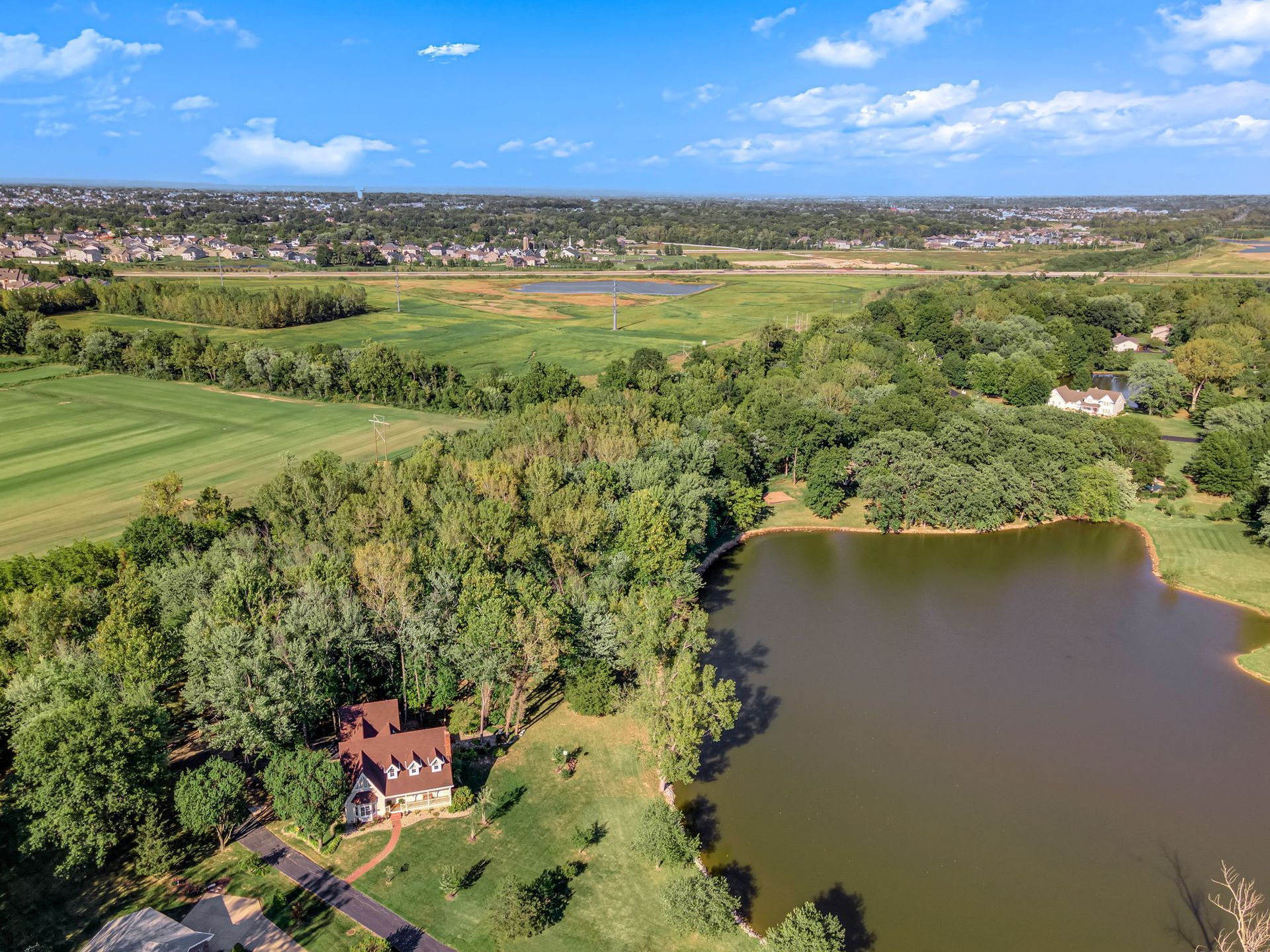 Aerial view of a red-roofed house beside a pond, surrounded by trees and green fields under a blue sky