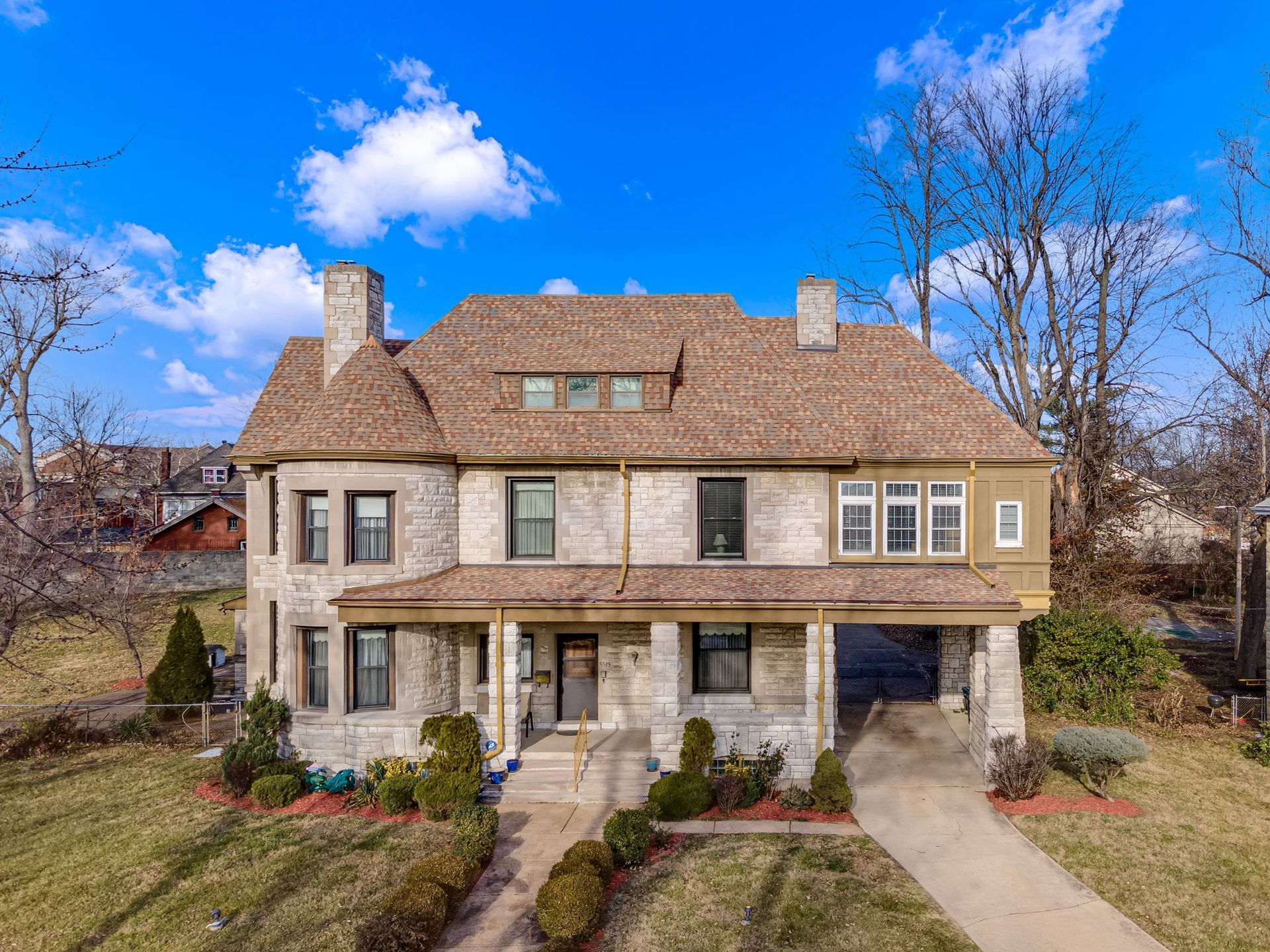 Large tan two-story house with a stone porch, steep shingled roof, and a partly cloudy blue sky