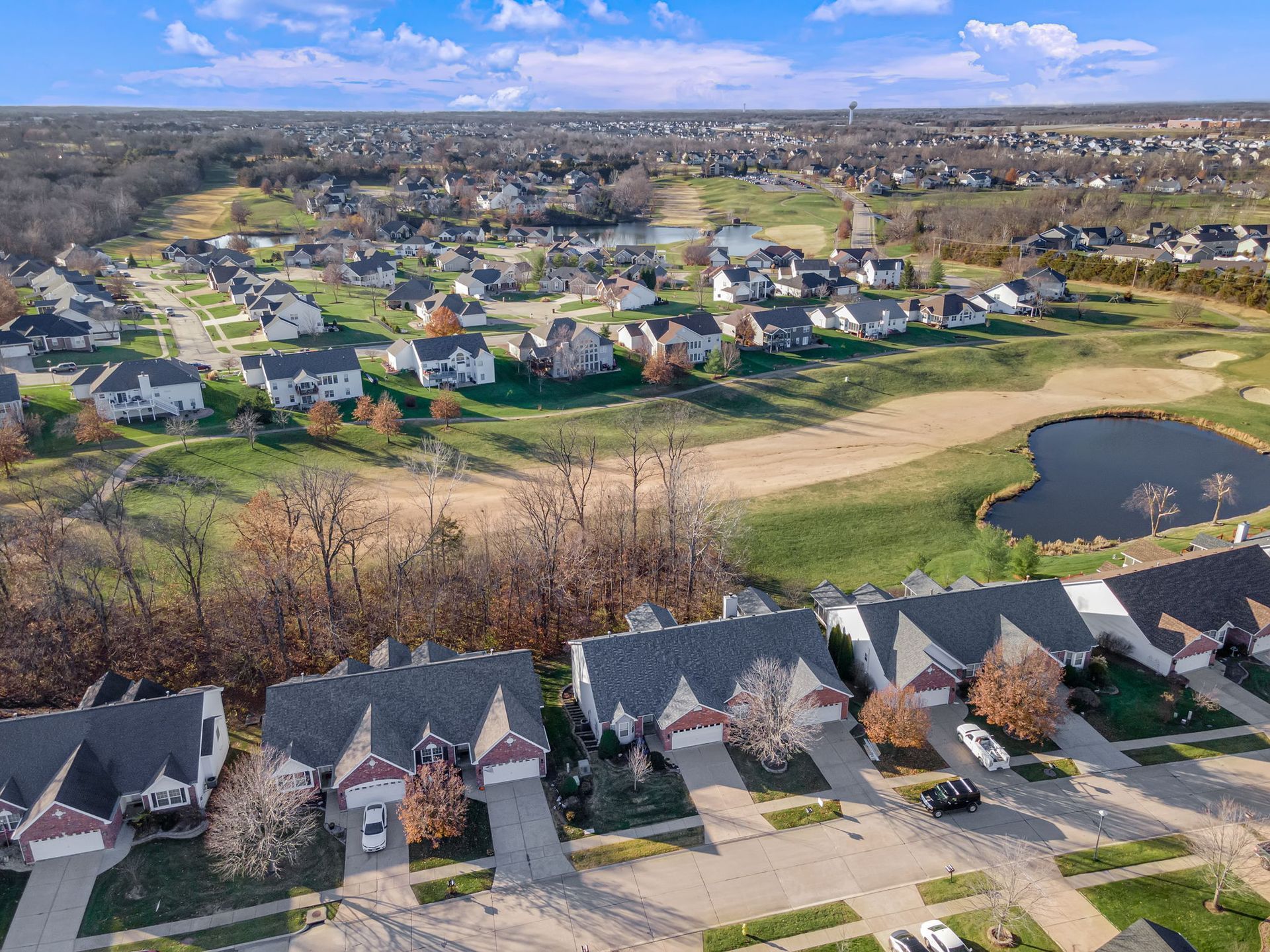 Aerial view of a suburban neighborhood with houses, green fields, and a small pond under a blue sky