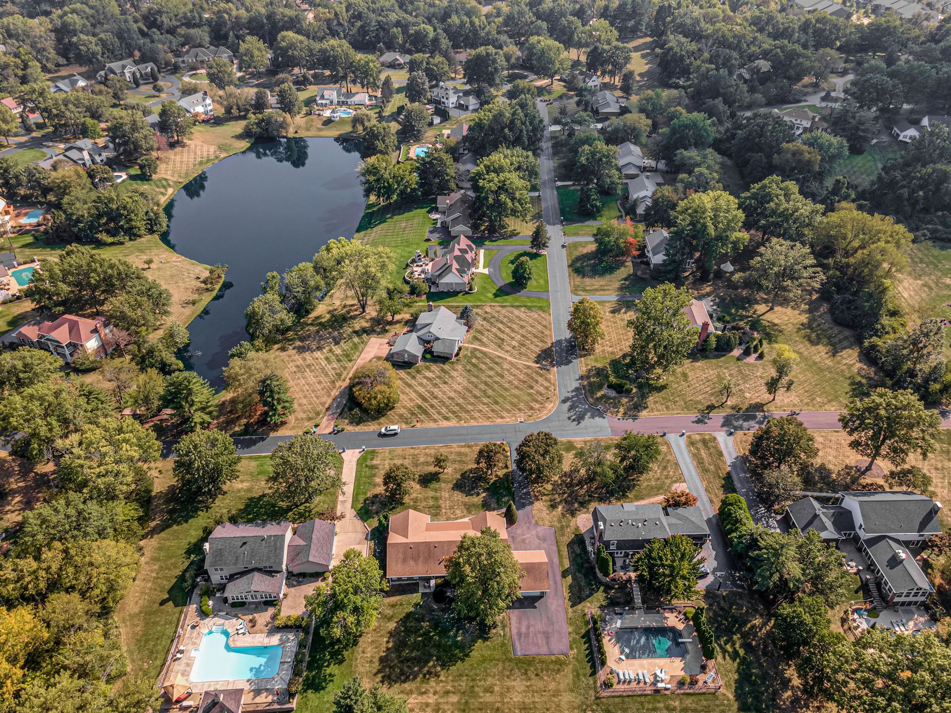 Aerial view of a suburban neighborhood with houses, swimming pools, trees, and a small pond.