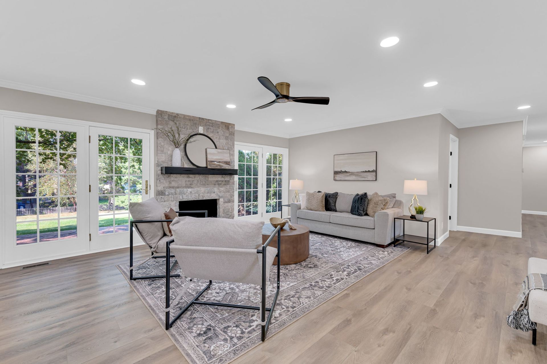 Bright open living room with stone fireplace, gray sofas, dining table, and large windows overlooking a pool.