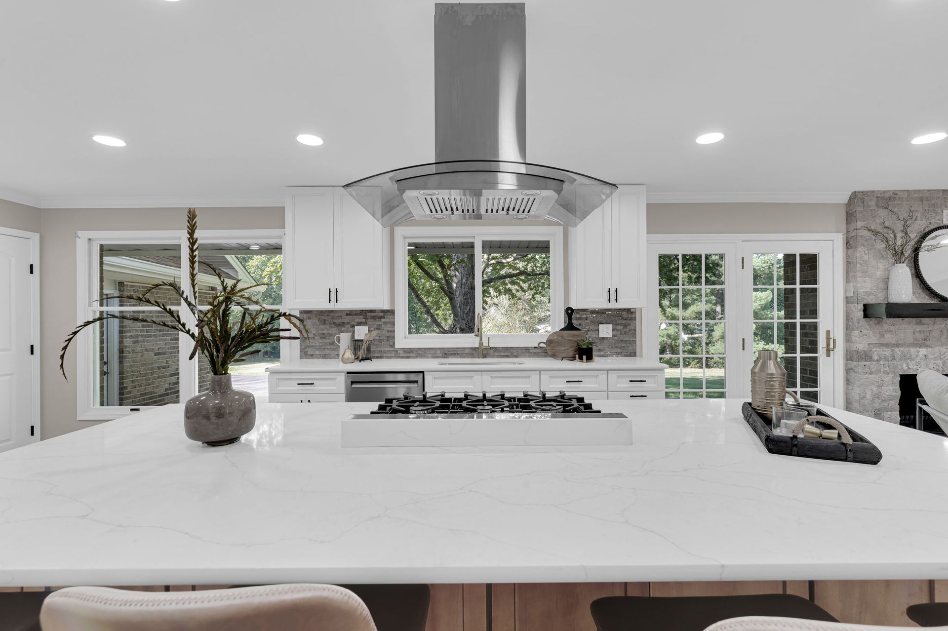 Bright modern kitchen with white island, stainless steel range hood, and large windows.