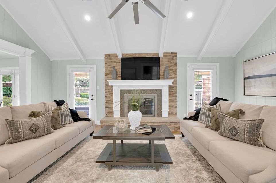 Bright living room with two beige sofas, a stone fireplace, white vaulted ceiling, and large windows