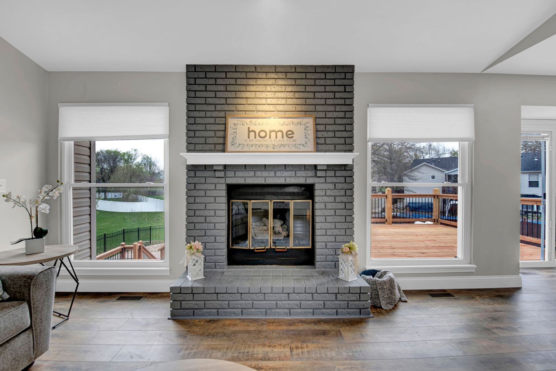 Bright living room with a brick fireplace, “home” sign, and large windows overlooking a deck.