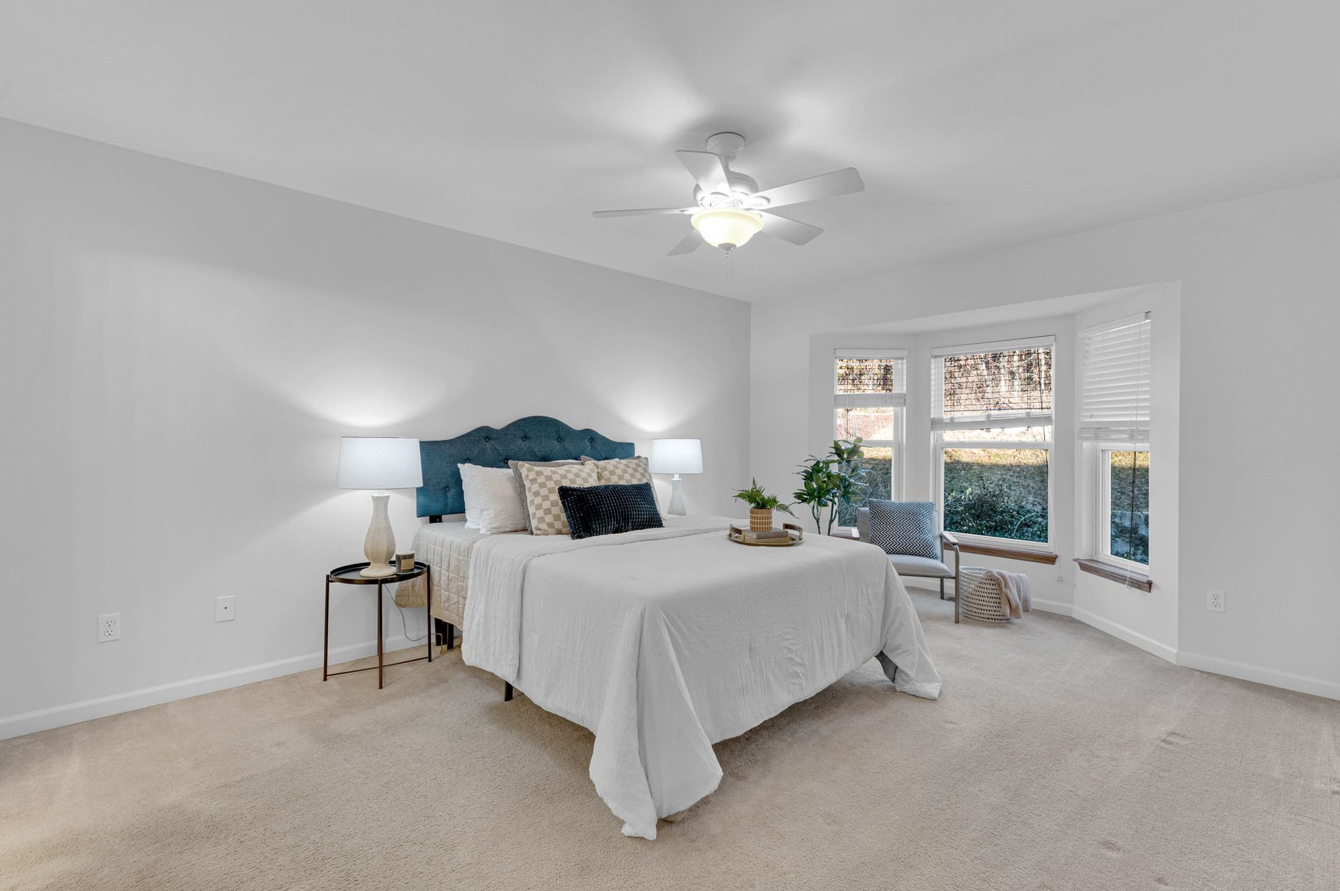 Bright bedroom with white bedding, blue headboard, nightstands, ceiling fan, and large windows with shutters