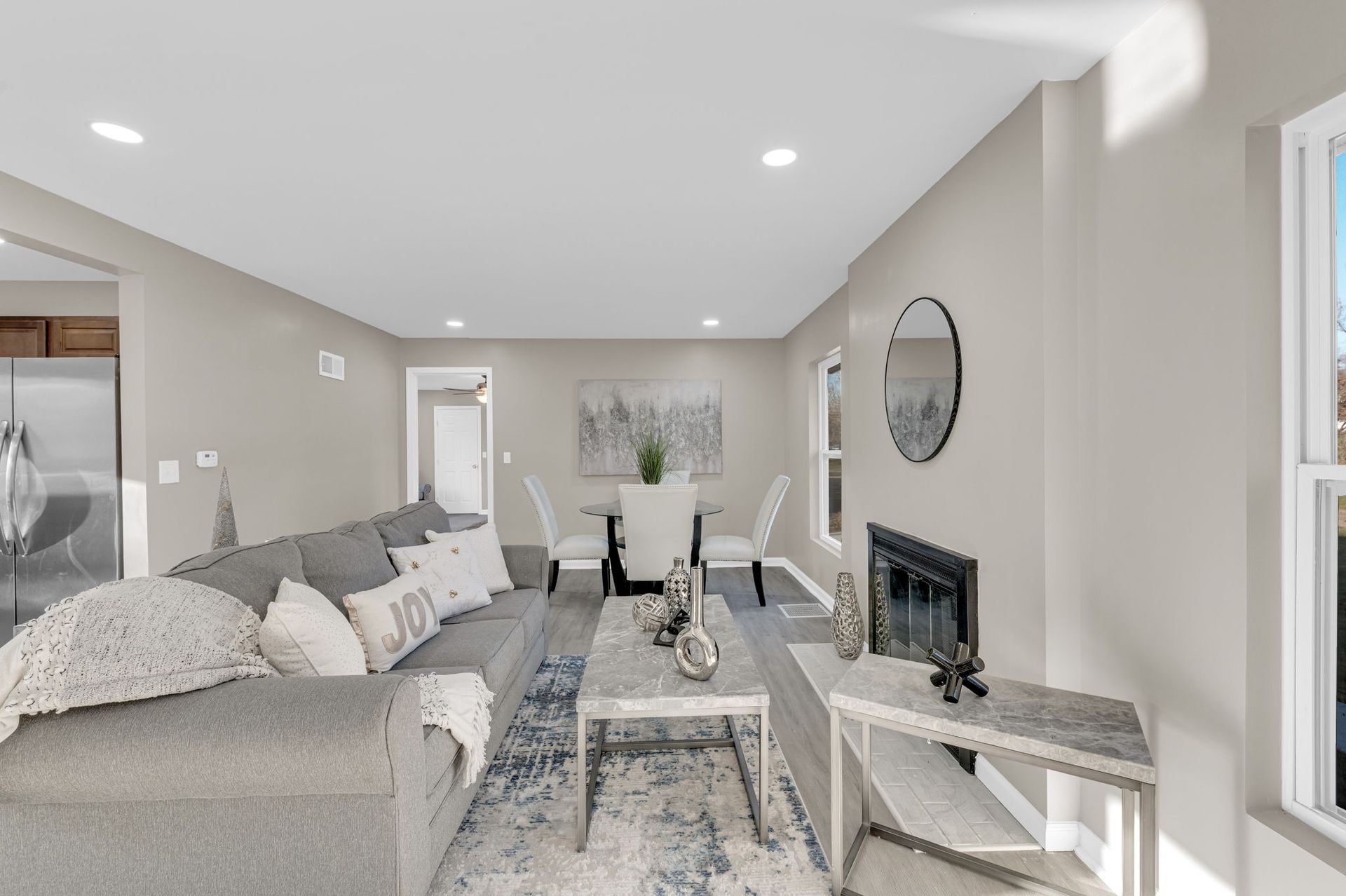 Bright living room with a gray sofa, wall-mounted fireplace, round mirror, and dining table by large windows