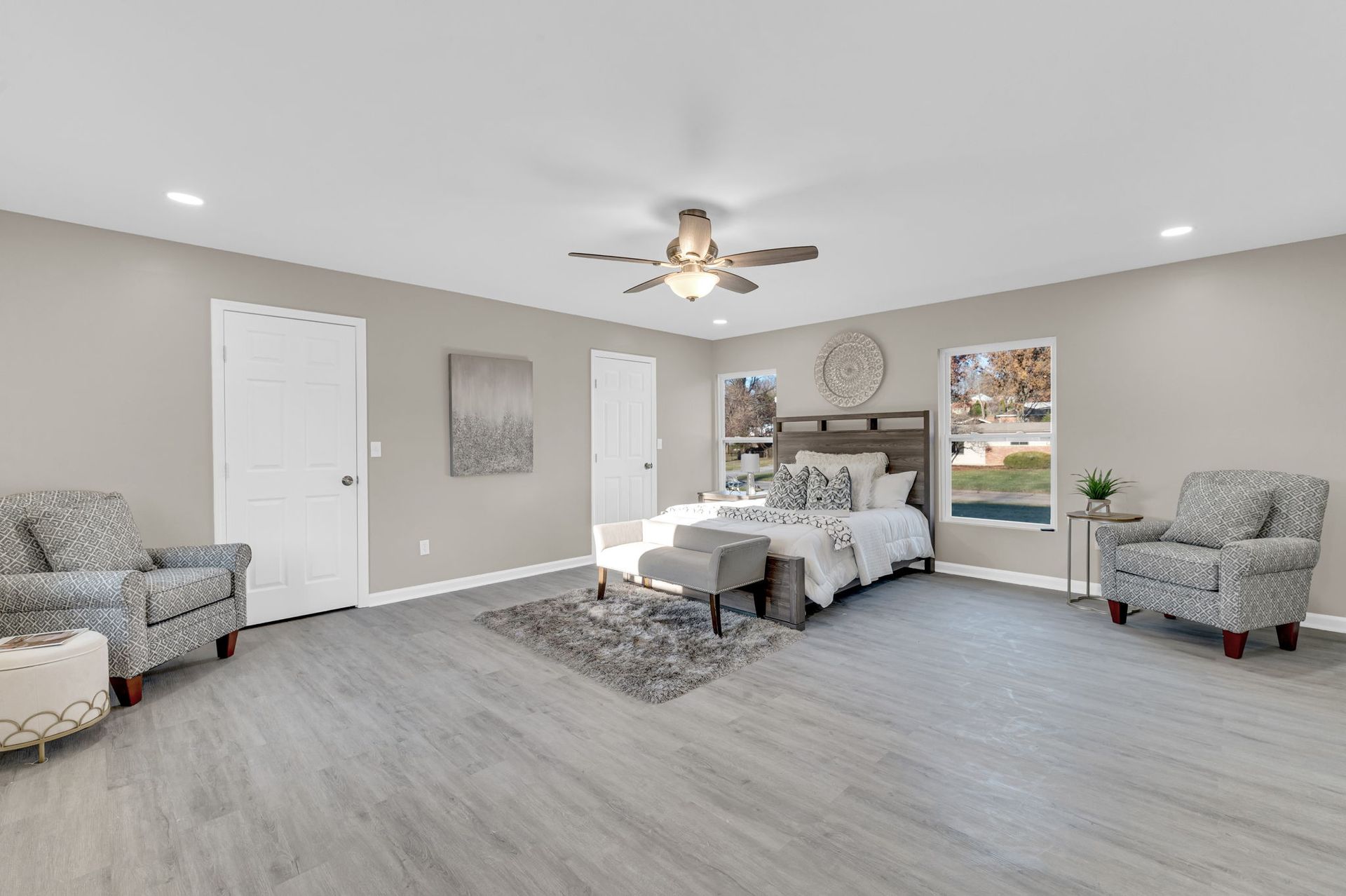 Spacious modern bedroom with gray walls, white bed, chairs, ceiling fan, and light wood flooring