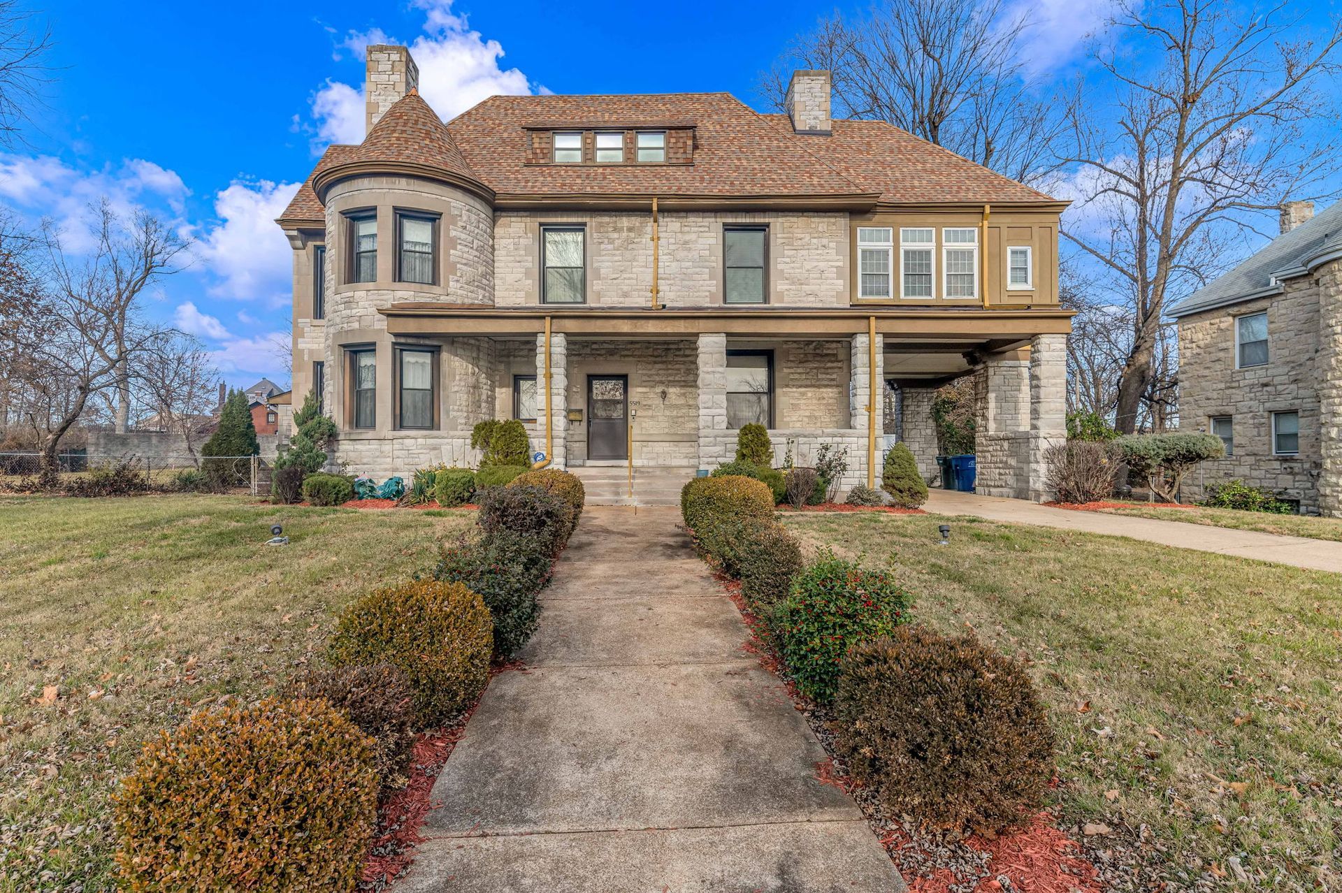 Large two-story stone house with a front walkway, arched porch, and manicured lawn under a blue sky