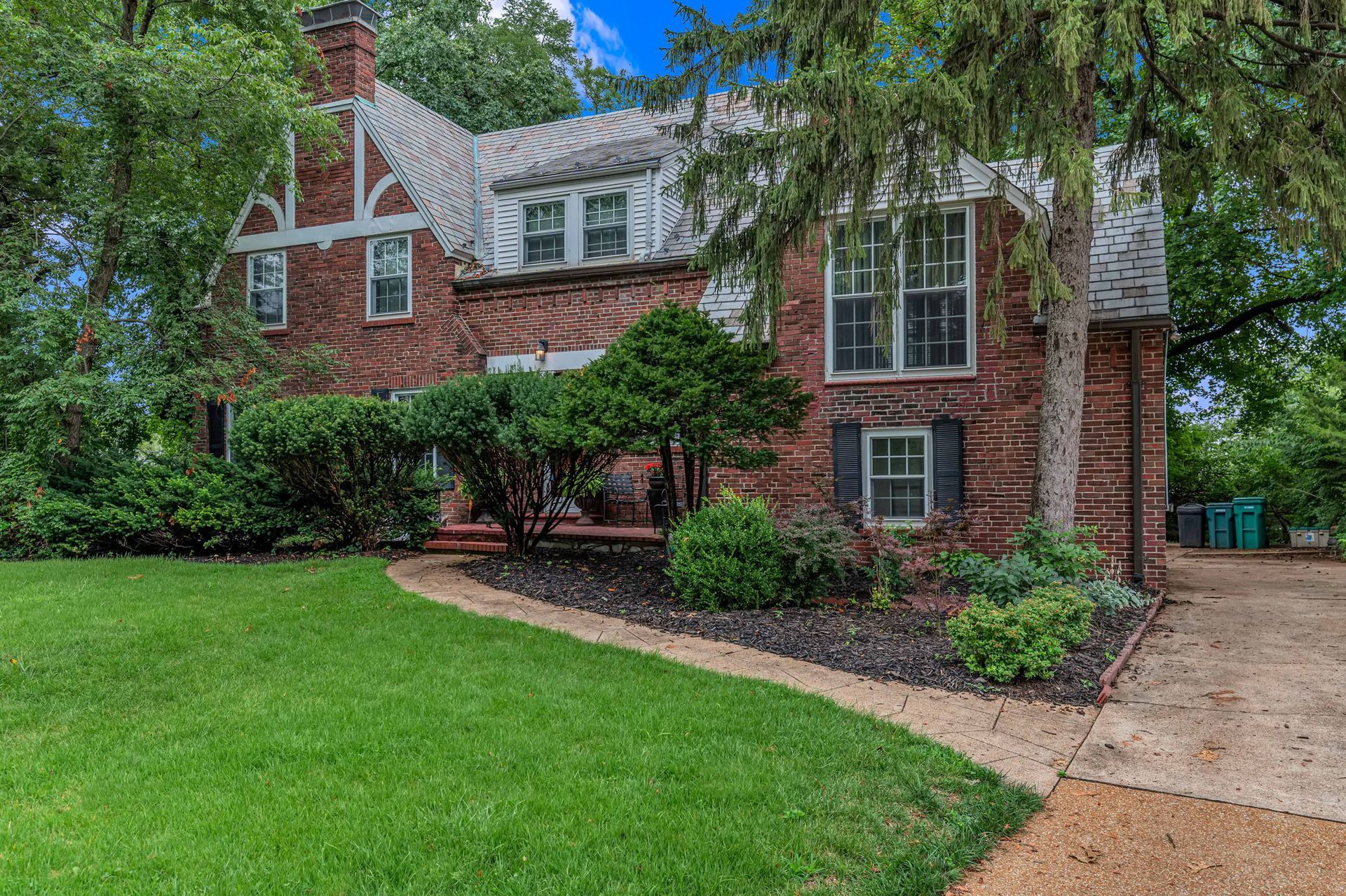 Brick house with white-trimmed windows, green lawn, and driveway surrounded by trees and shrubs