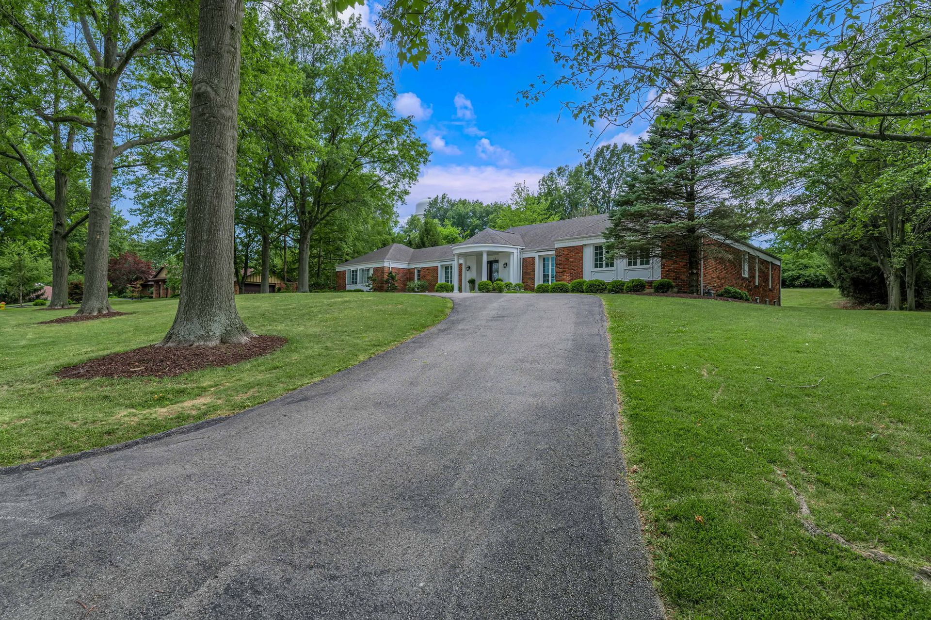Brick house with white columns at the end of a gravel driveway, surrounded by trees and green lawn