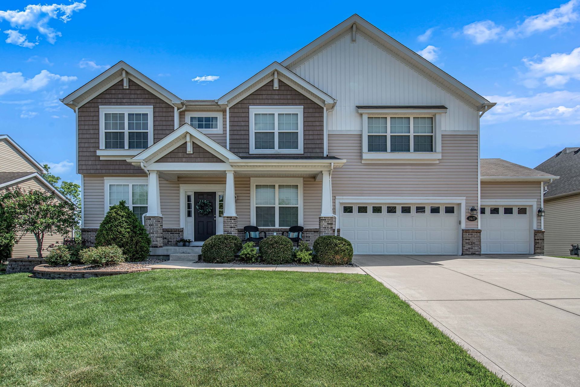 Two-story beige suburban house with a two-car garage, front lawn, and blue sky.