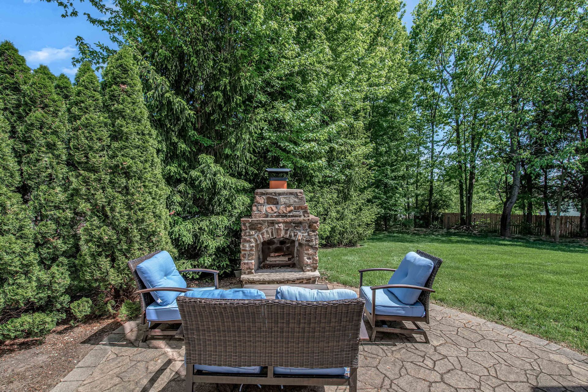 Backyard patio with wicker chairs around a brick outdoor fireplace, surrounded by trees and grass