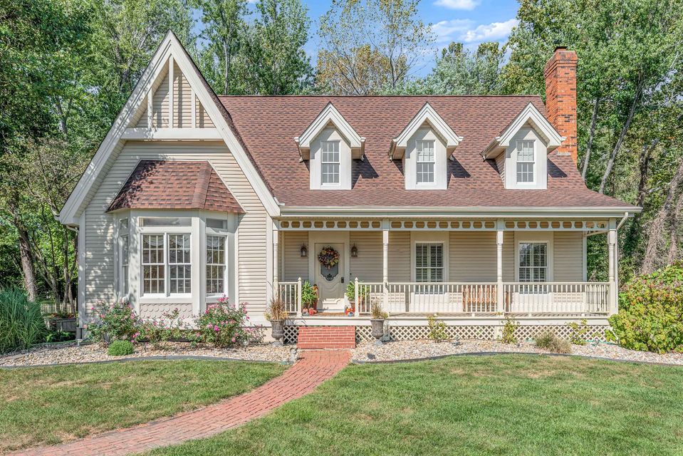Charming white cottage with red roof, front porch, dormer windows, and a brick path amid greenery.