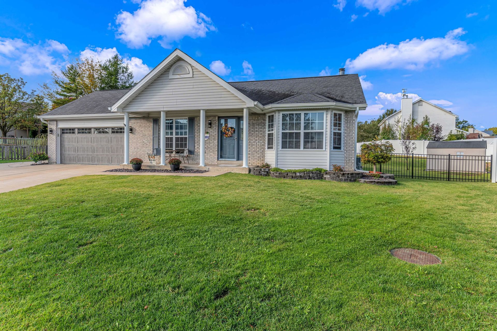 Single-story suburban house with a front porch, attached garage, and a large green lawn under a blue sky