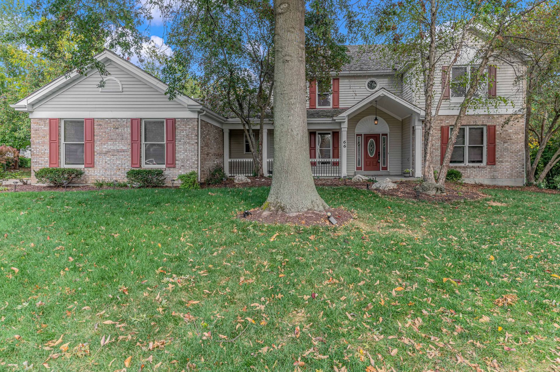Suburban brick house with red shutters, a front porch, and a large tree in the yard