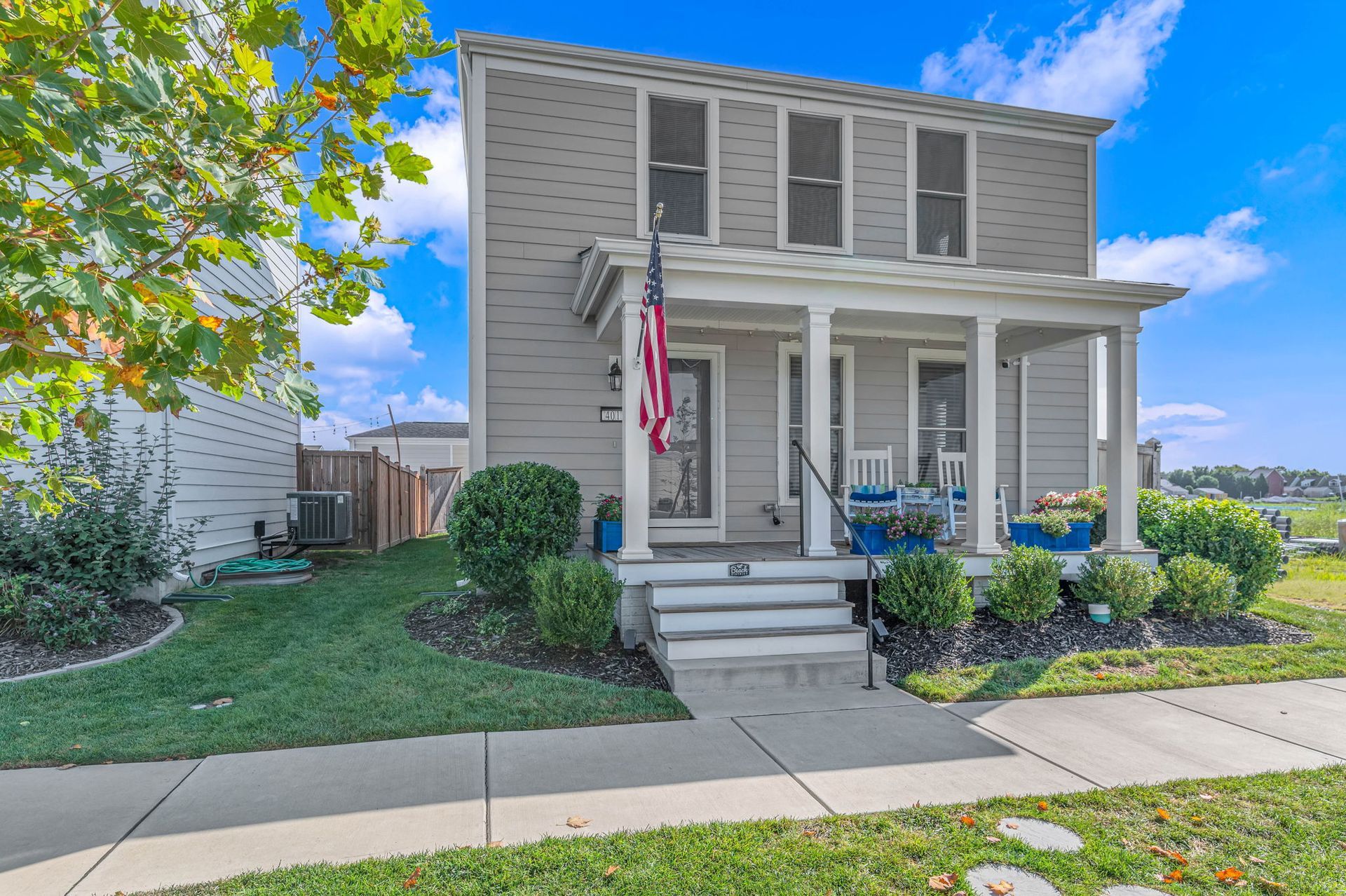 Two-story gray house with a front porch, American flag, and well-kept lawn on a sunny day