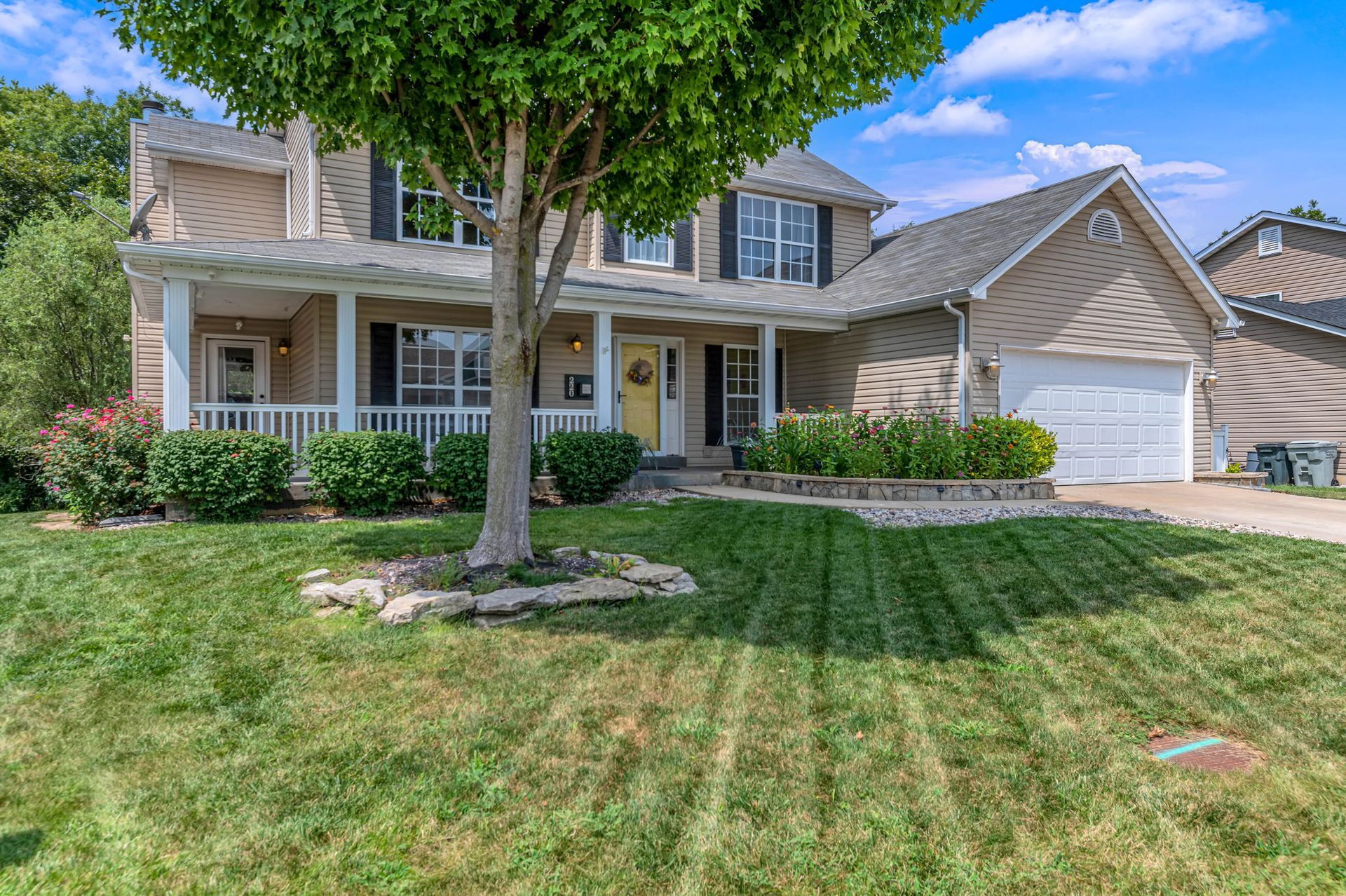 Two-story beige house with a front porch, white trim, and a large green lawn under a blue sky