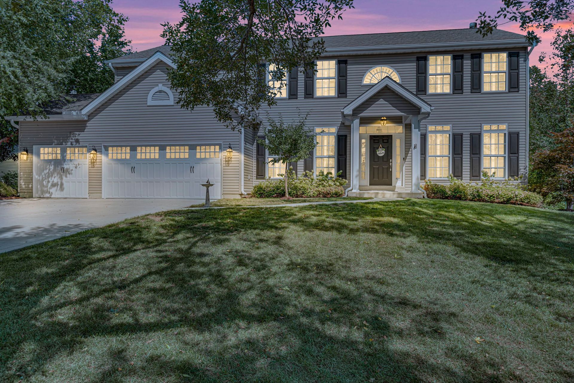 Two-story gray house with lit windows and garage at dusk, viewed from a grassy front yard.