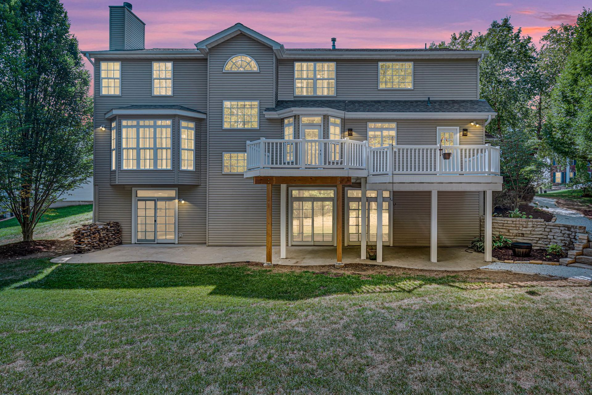 Large gray two-story house with a wraparound deck and lit windows at dusk in a wooded yard