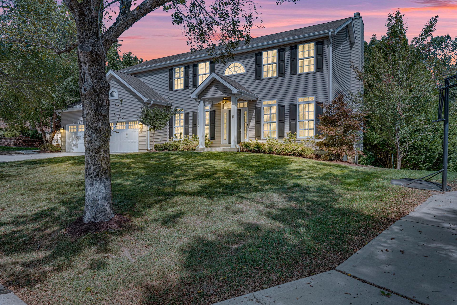 Two-story house with lit windows at dusk, a front lawn and tree in the yard, viewed from the sidewalk
