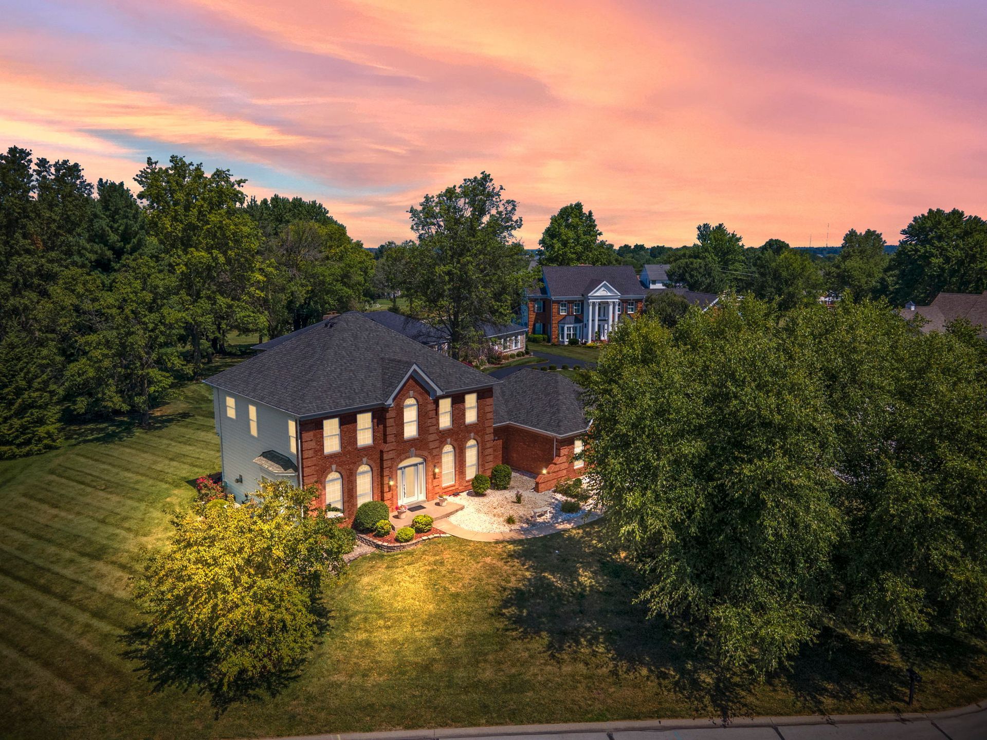 Large brick house with lit windows and patio at sunset, surrounded by trees and a grassy yard