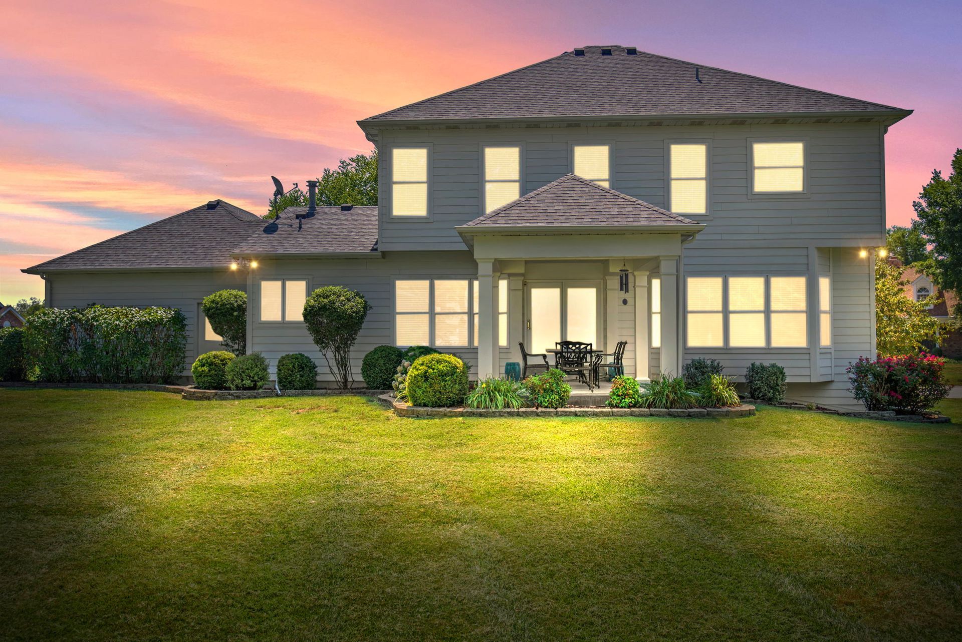 Large suburban house at sunset with lit windows, front porch, and a green lawn under a pink sky