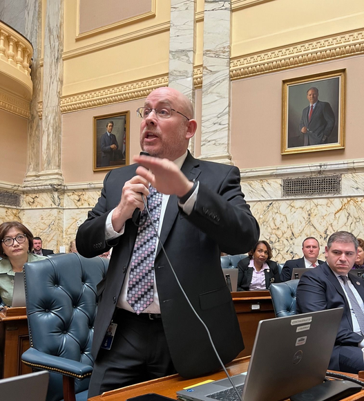 David speaking at a lectern in the state capital building, gesturing beside a laptop and seated attendees