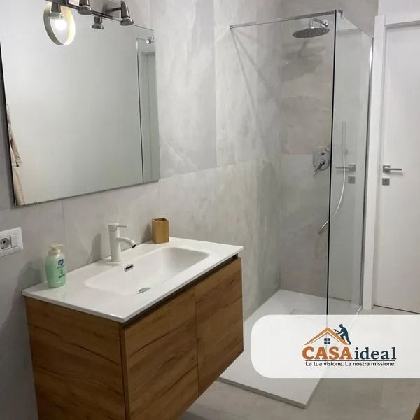 Bagno moderno con mobiletto in legno, lavabo bianco, ampio specchio, doccia in vetro e pareti piastrellate.