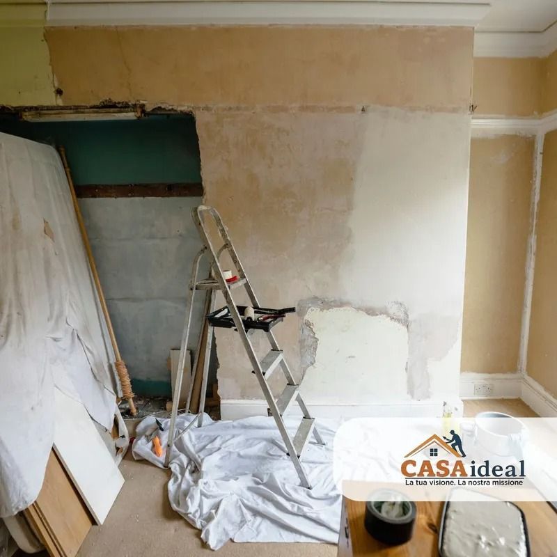 Stanza in fase di ristrutturazione con scala, polvere di intonaco e pareti beige parzialmente scrostate.