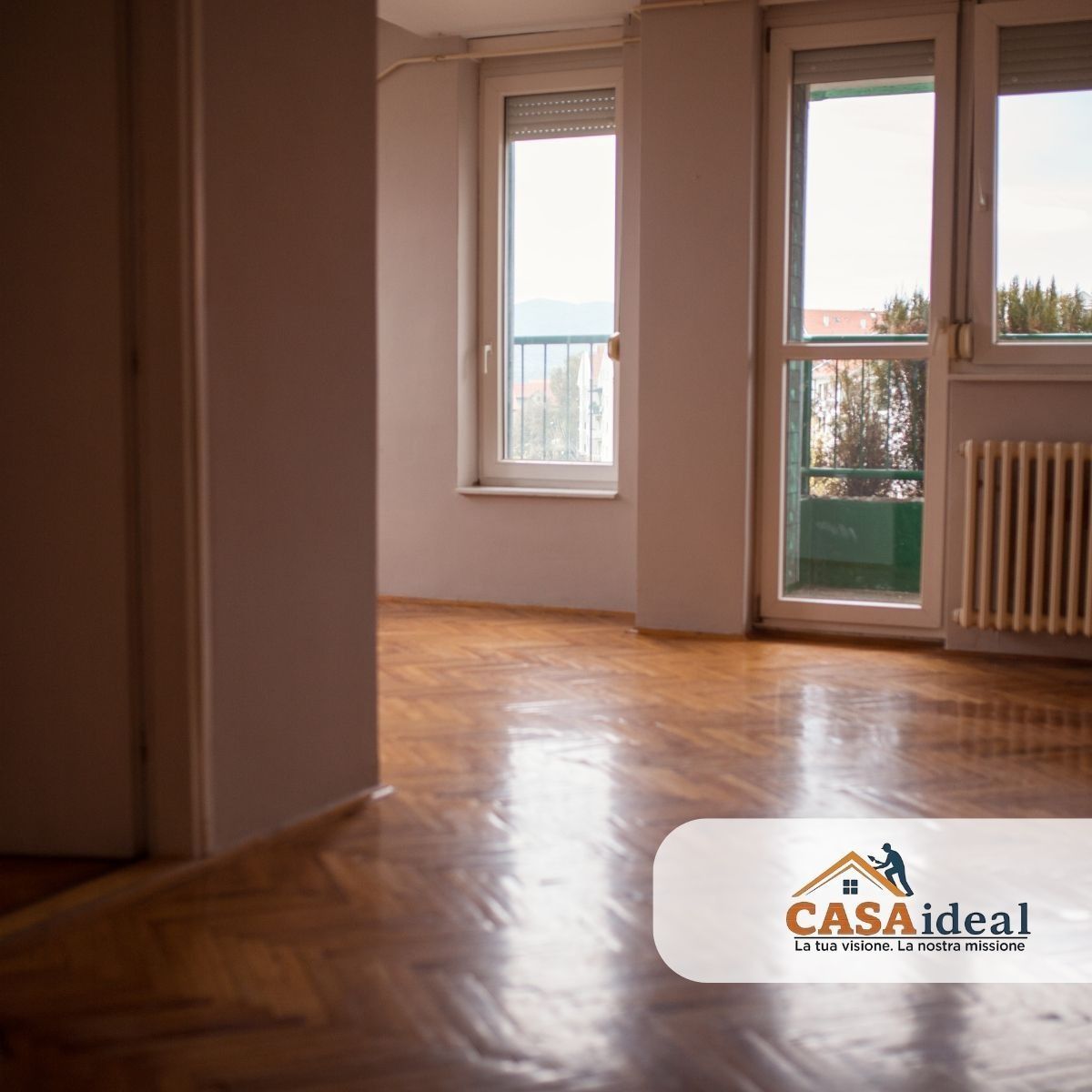 Stanza vuota e luminosa con pavimento in legno, finestre, porta-finestra e termosifone; logo CASAideal in un angolo.