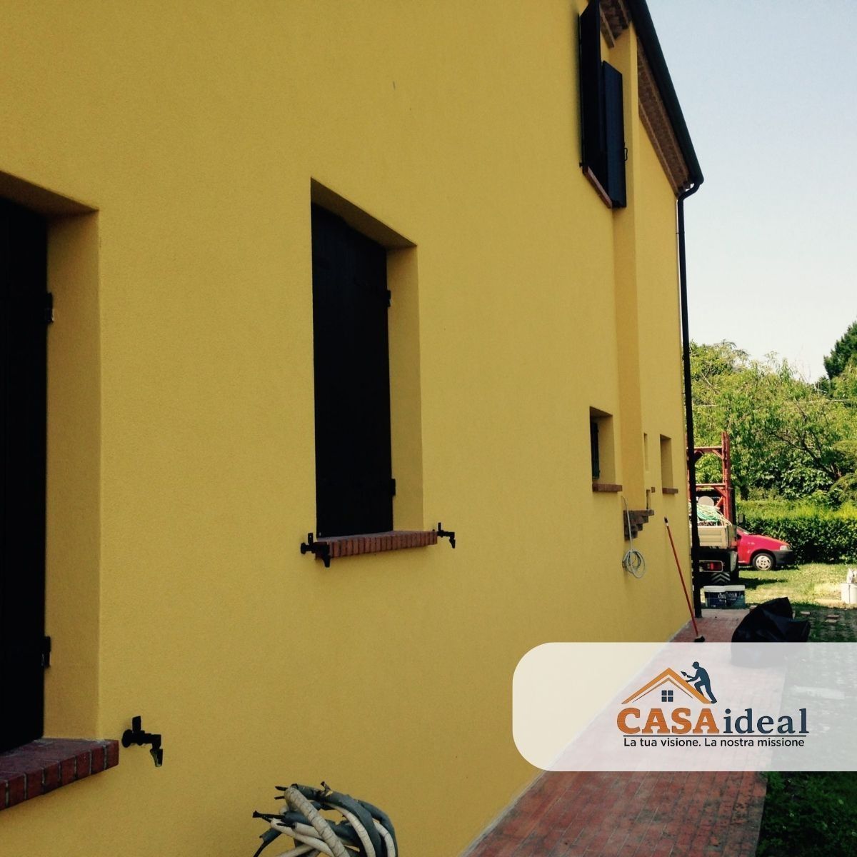 Esterno di una casa gialla con finestre scure, davanzali in mattoni e un piccolo giardino laterale.