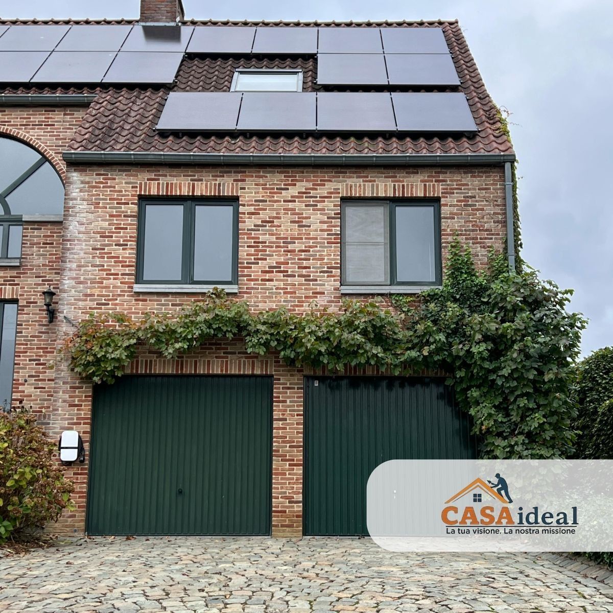 Casa a schiera in mattoni con porte del garage verdi, pannelli solari sul tetto scuro ed edera lungo la facciata principale.