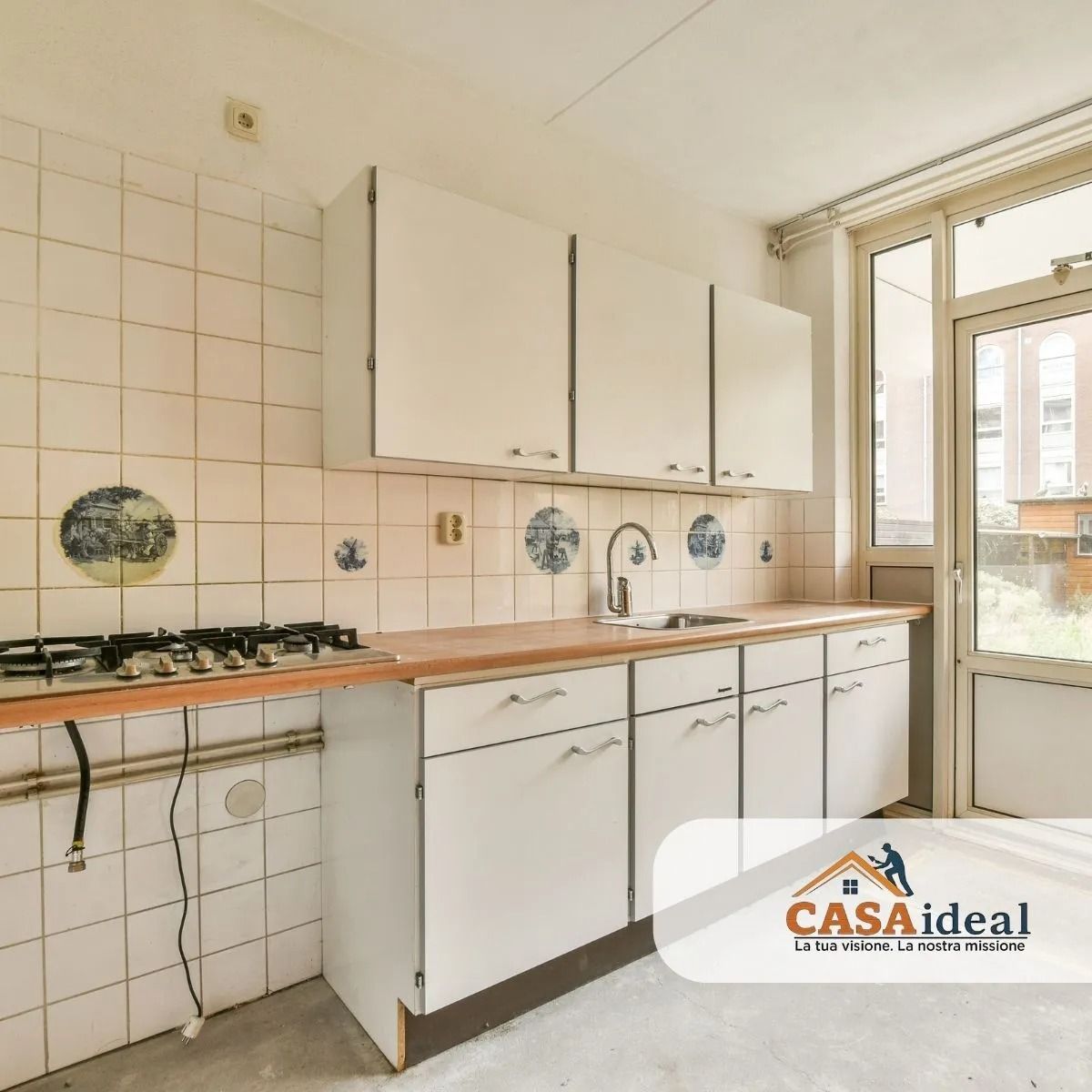 Piccola cucina bianca con pareti piastrellate, piano di lavoro in legno, fornello a gas e porta a vetri che dà su un balcone.