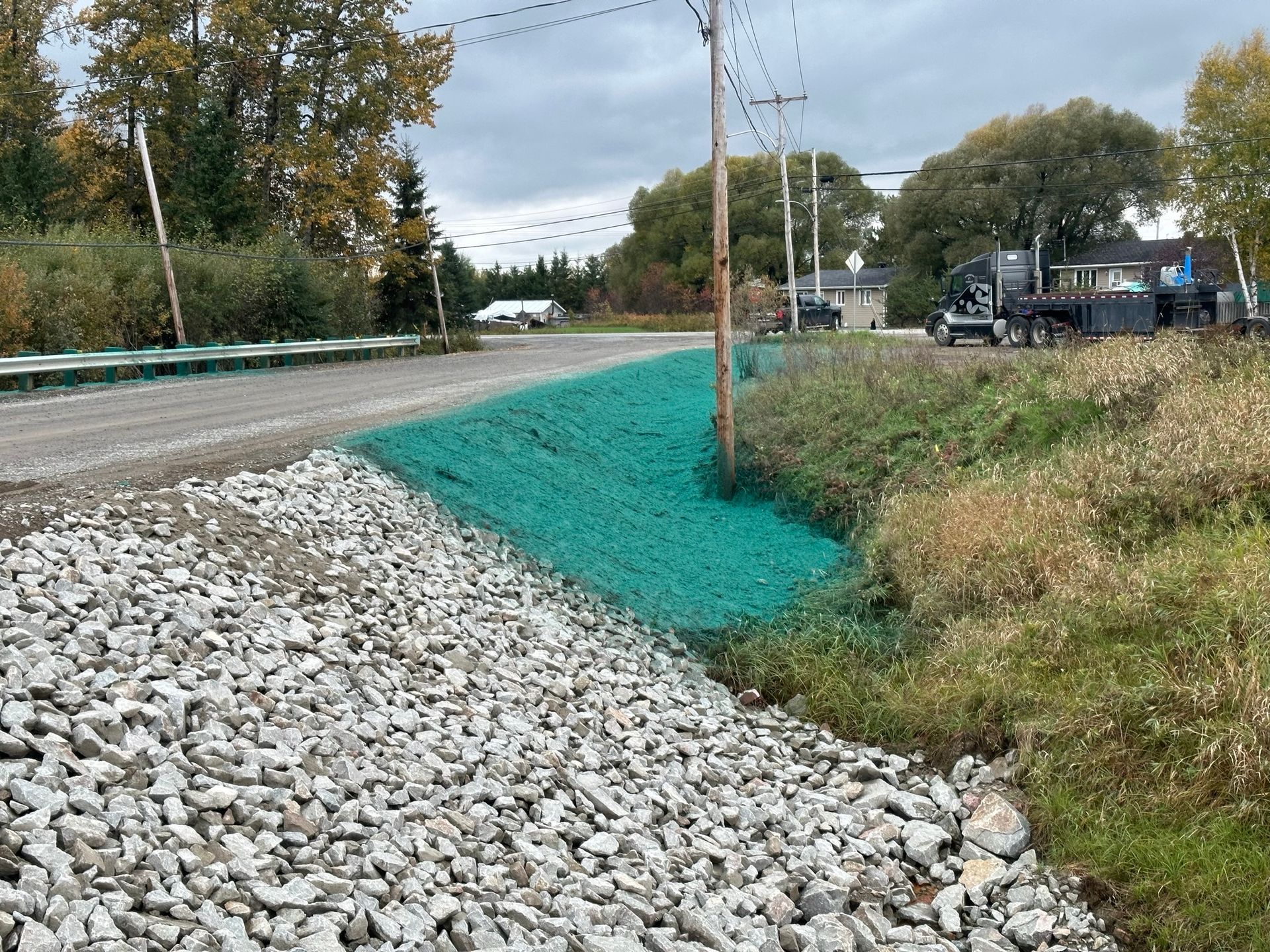 Travaux de construction en bord de route avec contrôle de l'érosion par voie verte sur une pente herbeuse, du gravier et un poteau électrique.