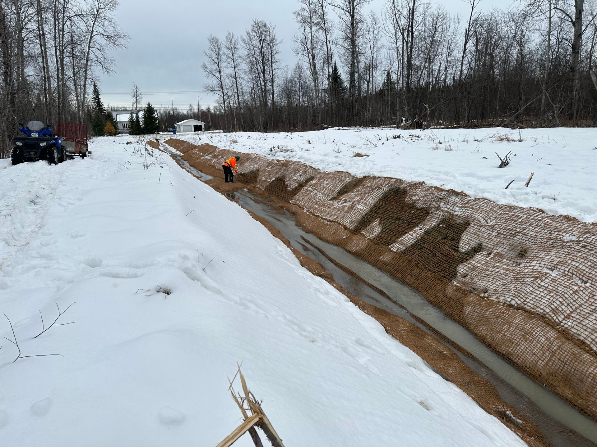 Un paysage enneigé avec une tranchée creusée en son centre. Une personne en gilet orange travaille près de la tranchée.