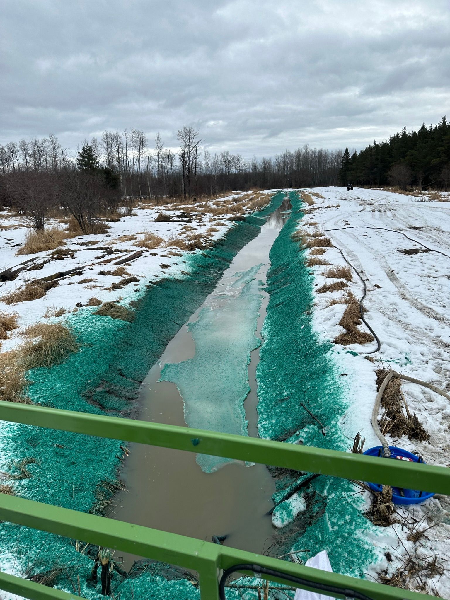 Un filet vert recouvre un fossé boueux dans un champ enneigé, vu depuis un pont verdoyant.