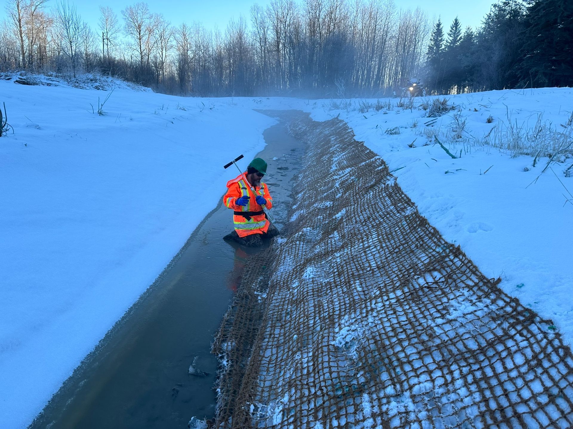 Une personne en tenue de travail orange est agenouillée dans un fossé enneigé, inspectant un tapis anti-érosion ; la forêt est visible en arrière-plan.