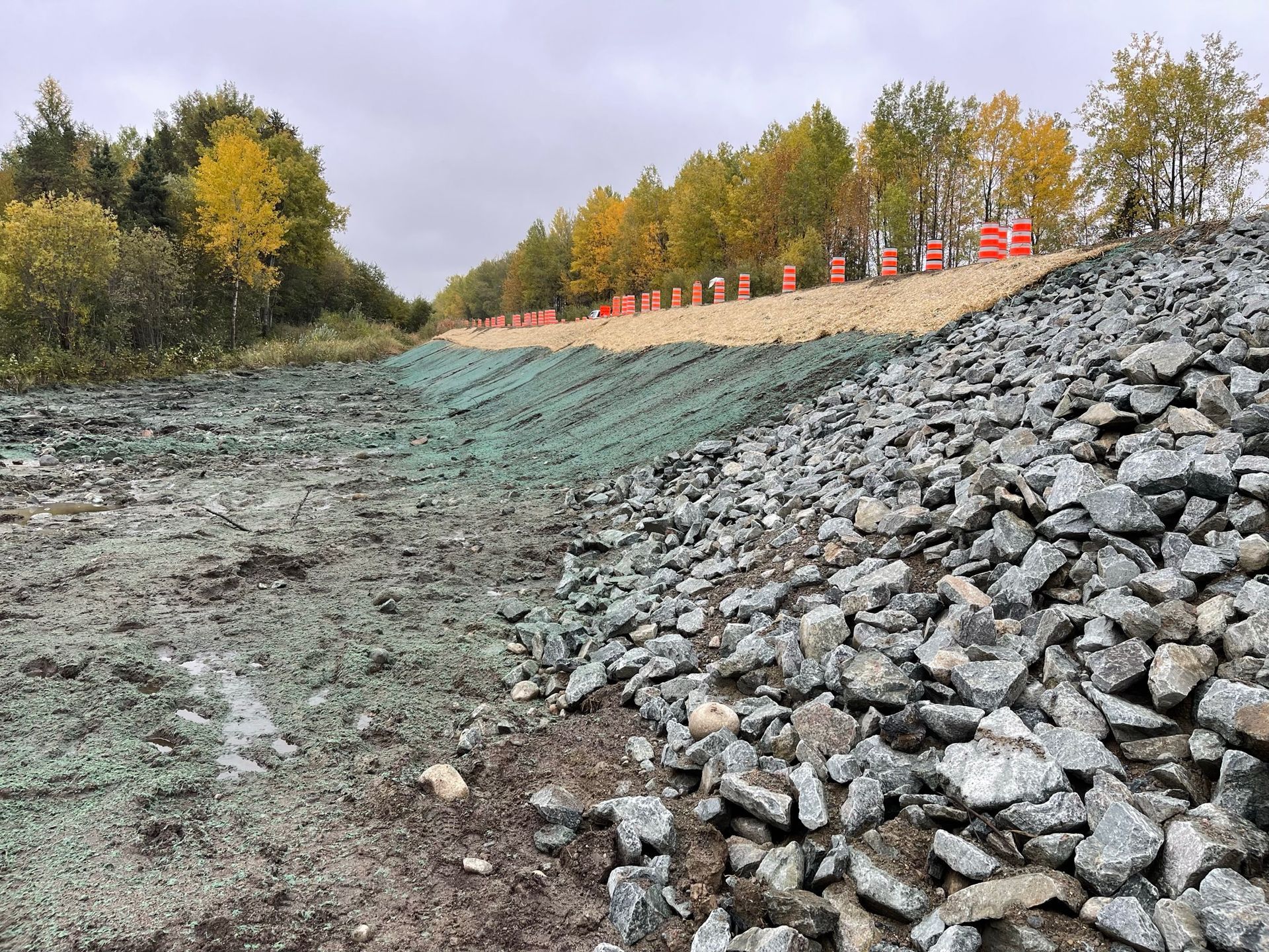 Chantier de construction avec gravier, filets verts et cônes orange à flanc de colline, parsemé de feuillages d'automne.