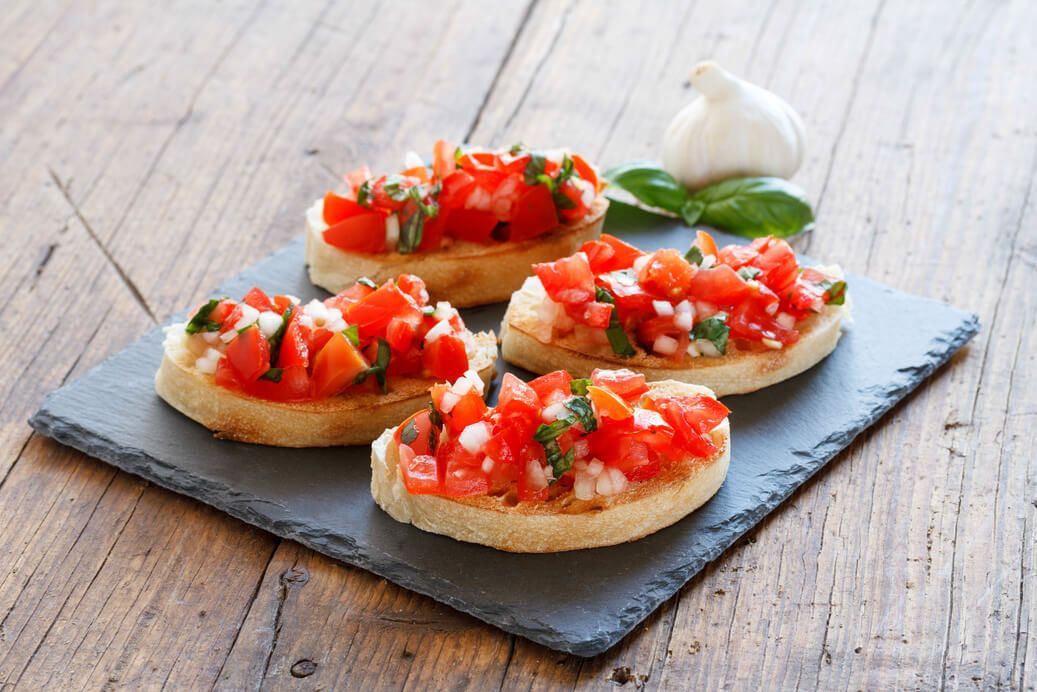 Quattro fette di pane condite con pomodori e basilico su un vassoio di ardesia su un tavolo di legno.