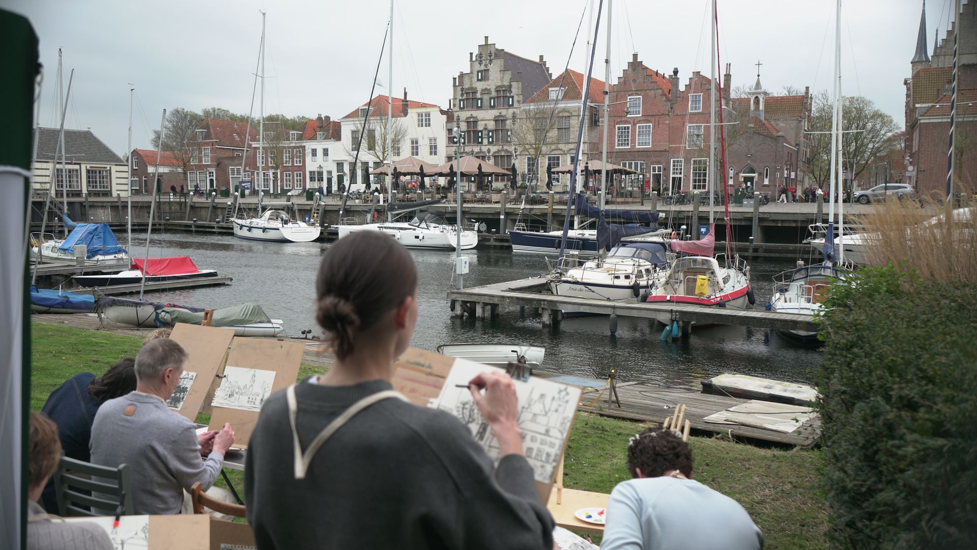 Mensen schetsen een havengezicht met boten en gebouwen langs een jachthaven. #schilderworkshop, schilderen, creatieve workshops in de regio