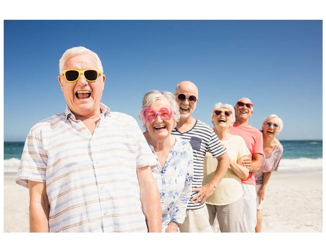 Group of smiling seniors wearing sunglasses at the beach, blue sky and ocean in the background.